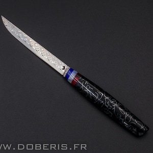 May include: A Damascus steel knife with a patterned blade and a black handle with a white design. The handle has a blue, white, and red accent. The knife is displayed on a black surface. The website address www.doberis.fr is at the bottom.