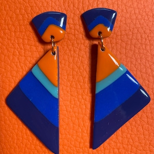 Peut inclure: Une paire de boucles d'oreilles géométriques au design rétro. Les boucles d'oreilles présentent un dessus bleu foncé, un connecteur doré et un pendentif triangulaire avec des rayures orange, bleues et turquoise. Les boucles d'oreilles sont posées sur un fond orange.