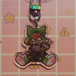May include: A clear acrylic keychain featuring a cartoon character with green and orange hair, wearing a black shirt with "SOUTH PARK" printed on it. The keychain has a rainbow-colored clasp and a pink background with repeating patterns.