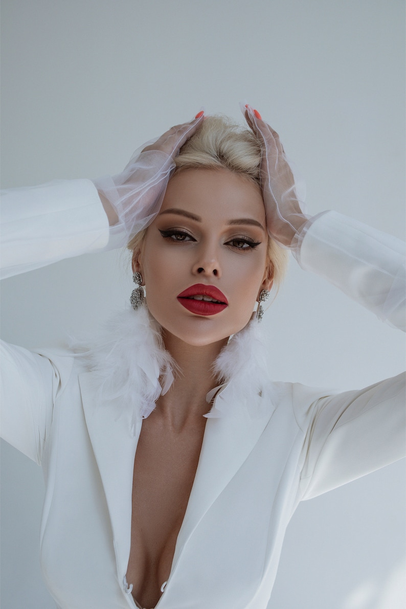 May include: A woman wearing a white blazer with a feather trim collar and white gloves. She has red lipstick and her hair is styled in a bouffant.