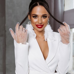 May include: A woman wearing a white blazer with a deep V-neckline and a white feather earring. She is wearing white mesh gloves and has her hair pulled back in a bun.