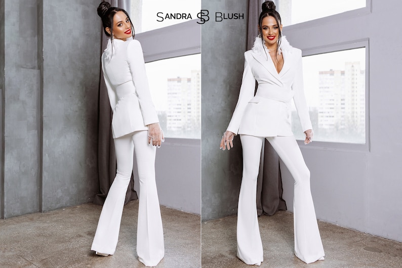 May include: A white pantsuit with a blazer and flared pants. The blazer has a notched collar and a double-breasted closure. The pants have a high waist and a wide leg. The model is wearing the pantsuit with a white fur collar.