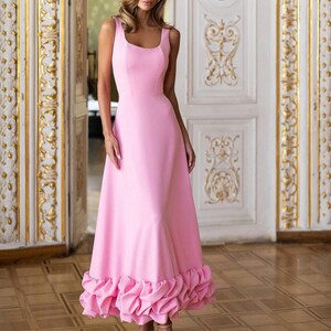 May include: A light pink, sleeveless dress with a square neckline and a flared skirt. The hemline has a ruffled design. The floor-length dress is worn with beige strappy heels. The background features ornate white doors with gold detailing.