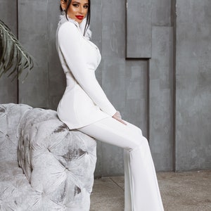 May include: A woman wearing a white pantsuit with a white feather collar sits on a white tufted sofa. She is looking at the camera and has red lipstick on.