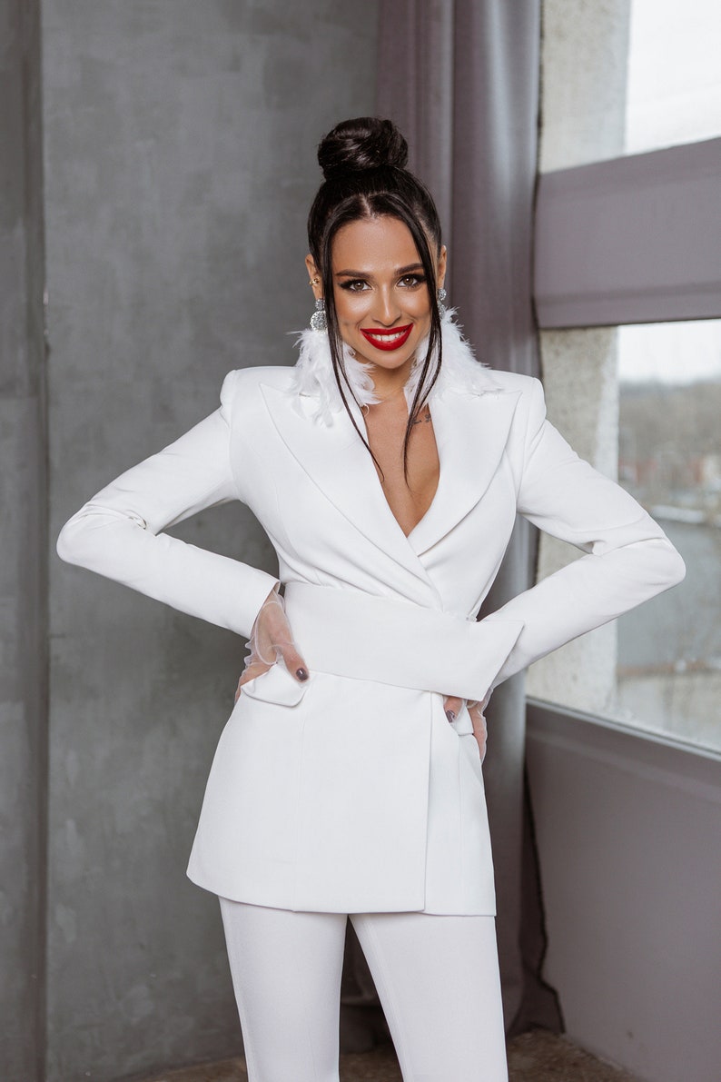 May include: A woman wearing a white suit with a deep V-neckline and a white feather trim around the collar. She is smiling and has her hands in her pockets.