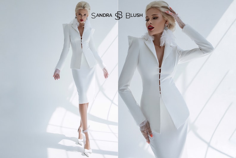 May include: A white tailored suit with a long-sleeved jacket and a knee-length skirt. The jacket has a pointed collar, a single-breasted closure, and a decorative feather trim. The suit is styled with white gloves and heels. The text "SANDRA B BLUSH" is visible in the image.