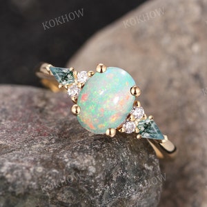 May include: A gold ring featuring an oval opal gemstone, flanked by two triangular blue-green stones and small white accent stones. The ring is set on a textured rock surface. The word "KOKHOW" is repeated in the background.