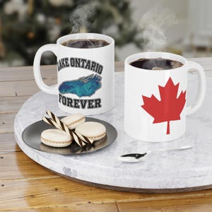 Peut inclure: Deux tasses en céramique blanche remplies d'un liquide sombre fumant. L'une présente un graphique bleu marbré du lac Ontario avec le texte "LAKE ONTARIO FOREVER" en noir. L'autre affiche une grande feuille d'érable rouge. Trois macarons sont sur une assiette.