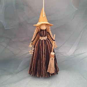 Puede incluir: Un adorno de muñeca colgante con un sombrero de bruja de fieltro beige, cabello de lana rubia larga y ondulada, y un vestido hecho de hilos de lana negros y marrones. La figura sostiene una escoba pequeña con un dije de corazón y tres estrellas.