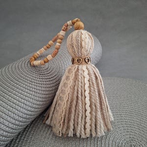 Peut inclure: Un pompon en laine beige et marron avec une perle en bois et des breloques en forme de cœur. Le pompon est attaché à une chaîne de perles en bois.