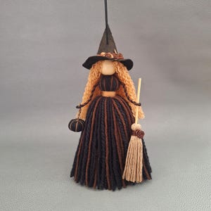 May include: A hanging witch ornament crafted from yarn, featuring long curly blonde hair, a black pointed hat, and a dress made of alternating black and brown yarn strands. The figure holds a small black pumpkin and a miniature tan broomstick.