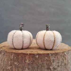 May include: Two white felt pumpkins with silver and gold trim. The pumpkins are sitting on a wooden surface.