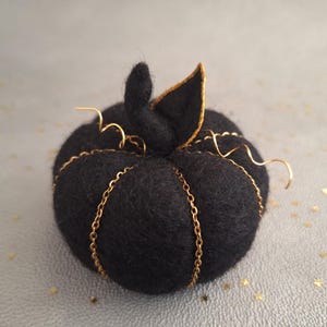 Könnte beinhalten: Ein schwarzer Filz-Kürbis mit einer goldenen Kette darum gewickelt. Der Kürbis hat einen kleinen Filzstiel mit goldener Umrandung.