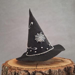 Pode incluir: Um chapéu de bruxa preto com uma teia de aranha e um amuleto de aranha, alfinetes de segurança e uma corrente prateada com contas prateadas.
