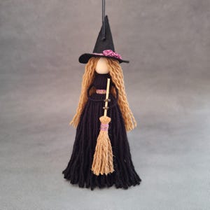 Könnte beinhalten: Eine schwarze Hexenpuppe mit langen braunen Garnhaaren, die einen schwarzen Filzhut mit rosa Blumen trägt und einen braunen Garnbesen hält.