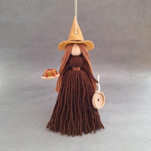 May include: A brown yarn doll with long brown hair wearing a brown felt witch hat and holding a plate of pancakes. The doll is wearing a brown yarn dress and a brown belt.
