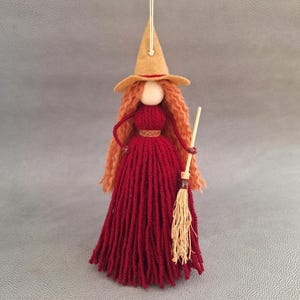 Puede incluir: Una muñeca de bruja de hilo rojo con un sombrero de fieltro marrón y pelo largo de hilo naranja sosteniendo una pequeña escoba.