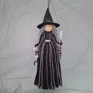 May include: A hanging doll ornament featuring a black pointed hat, long gray yarn hair, and a dress made of alternating black and gray yarn strands. The figure holds a black wand with white spider accents and a clear crystal-like topper.