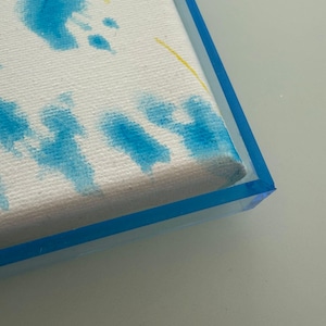 May include: A close-up of a canvas with blue and yellow paint splatters. The canvas is framed in a clear, blue-edged acrylic frame. The artwork has a textured surface.