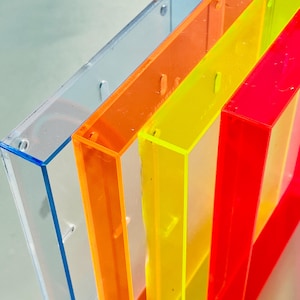 Pode incluir: Uma fileira de quatro molduras retangulares de acrílico transparente em cores vibrantes: azul claro, laranja, amarelo e vermelho. Cada moldura tem um interior oco e orifícios para parafusos visíveis, sugerindo que são projetadas para conter fotos ou obras de arte. As molduras estão em ângulo.