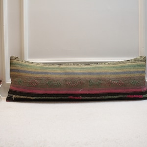 Puede incluir: Una almohada larga y rectangular con un patrón de rayas coloridas. La almohada está hecha de tela tejida con un borde marrón oscuro. La almohada está sobre una superficie blanca.