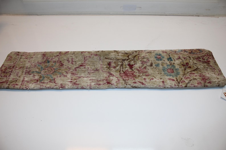 May include: A rectangular, vintage-style pillow with a faded floral pattern in muted tones. The design features burgundy, teal, and olive green against a beige background, creating a distressed, antique look. The pillow is likely for home decor.