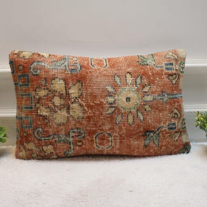 Puede incluir: Cojín rectangular de estilo vintage con un aspecto desgastado y texturizado. El cojín presenta un fondo naranja rojizo cálido con patrones florales y geométricos descoloridos en beige, azul y verde. Un elemento decorativo para el hogar.