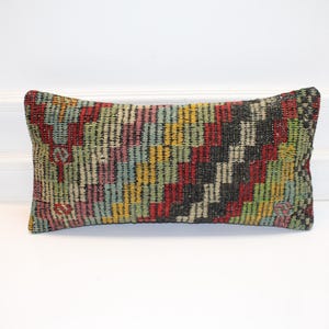 May include: Rectangular decorative pillow with a woven, geometric pattern. The pillow features a mix of colors including red, black, green, yellow, and blue. The pillow is placed on a white surface.