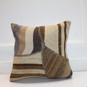 May include: Square decorative pillow with a patchwork design. The pillow features a mix of neutral colors, including beige, brown, and gray, arranged in vertical and diagonal stripes and geometric shapes. The pillow is made of a textured fabric.