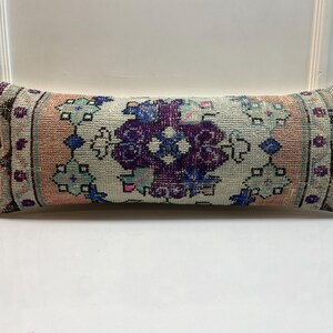 May include: A rectangular cushion with a vintage rug design. The cushion features a central floral pattern in purple and blue, framed by a light beige background and bordered by pink and teal accents. It is placed on a white surface.