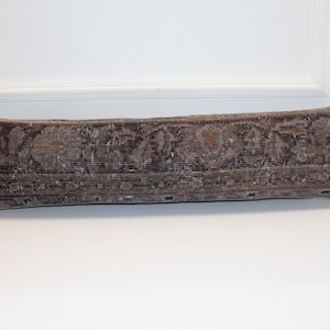 May include: A rectangular, brown, vintage-style pillow with a faded floral pattern. The pillow has a distressed look, with a mix of brown, gray, and beige tones. It is placed against a white background.