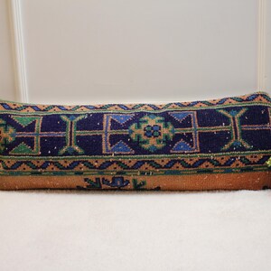 May include: A long, rectangular pillow with a colorful geometric pattern. The pillow is made of a woven fabric with a blue, green, orange, and white design. The pillow is lying on a white surface.