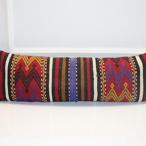 Puede incluir: Un cojín largo y rectangular con un diseño tejido. El cojín presenta un fondo rojo con rayas verticales negras, blancas, marrones y azules. El centro del cojín tiene un patrón geométrico repetitivo en rojo, amarillo y azul.