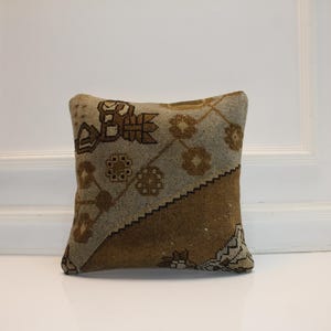 May include: Square decorative pillow with a geometric pattern in shades of brown, beige, and tan. The pillow has a textured appearance and is placed against a white background. The pillow's design includes floral and geometric shapes.