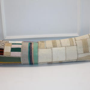 May include: A long, rectangular pillow with a patchwork design. The pillow features various fabric patches in shades of beige, green, pink, and brown. The pillow is approximately 36 inches long.