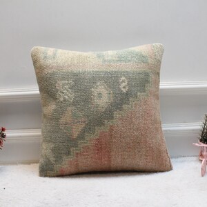 Puede incluir: Una almohada cuadrada con fondo beige y un patrón geométrico verde y rosa. La almohada está hecha de una tela suave y tejida.