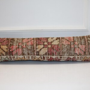 Op de afbeelding: Een lang, rechthoekig kussen met een geweven, patroonontwerp. Het kussen heeft een mix van kleuren, waaronder roze, beige, bruin en groen. Het patroon lijkt een bloemen- of geometrisch ontwerp te zijn. Het kussen ligt op een wit oppervlak.