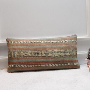 May include: A long rectangular pillow with a woven pattern in shades of beige, brown, and green. The pillow has a striped design with a decorative border.