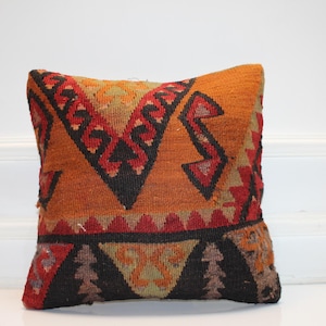 May include: A square, woven pillow with a geometric pattern in shades of orange, red, black, and tan. The pillow features a central design with zig-zag and triangular shapes, creating a textured, decorative accent.