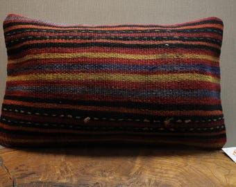 May include: Rectangular pillow with a woven, striped pattern in shades of red, orange, yellow, blue, and black. The pillow is placed on a wooden surface, and a small tag is visible.