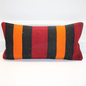 Puede incluir: Cojín rectangular con un patrón de rayas en negro, naranja y rojo. El cojín es un elemento decorativo, probablemente para un sofá o cama. Las rayas son verticales y están espaciadas uniformemente.