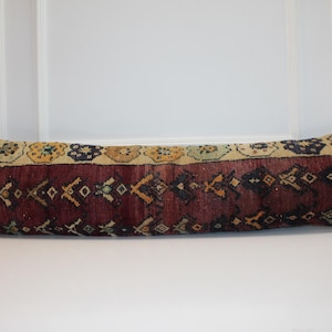 May include: A long, rectangular pillow with a patterned design. The pillow features a burgundy background with a repeating geometric pattern in gold, black, and cream. The top edge has a cream border with floral and geometric motifs.