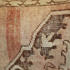May include: A woven textile cushion cover featuring a faded peach and beige color palette with a prominent dark brown geometric pattern. The fabric displays a visible knotted texture with a decorative border along the top edge of the piece.