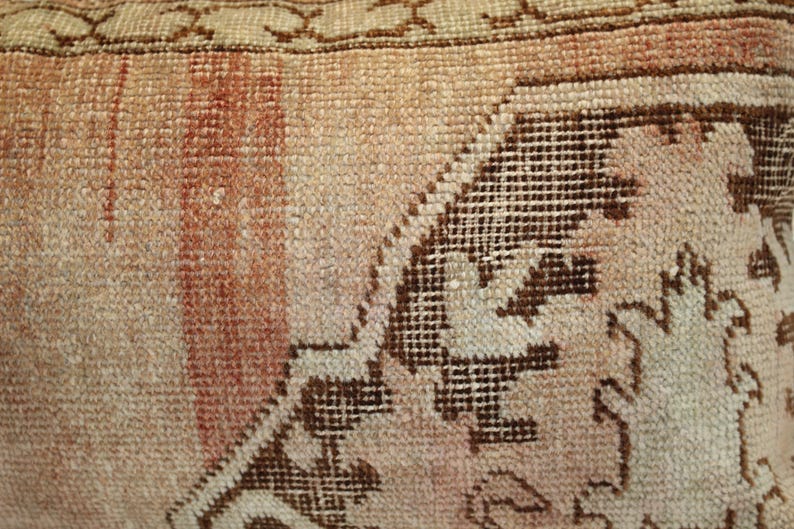 May include: A woven textile cushion cover featuring a faded peach and beige color palette with a prominent dark brown geometric pattern. The fabric displays a visible knotted texture with a decorative border along the top edge of the piece.