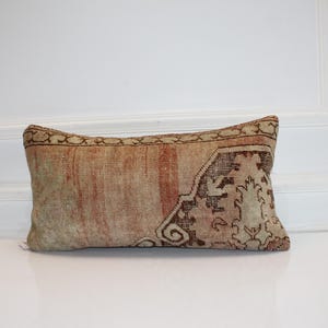 May include: A rectangular lumbar pillow featuring a faded, weathered textile cover with a muted color palette of beige, soft red, and brown. The fabric displays a worn, distressed texture with intricate, faded geometric and floral patterns.