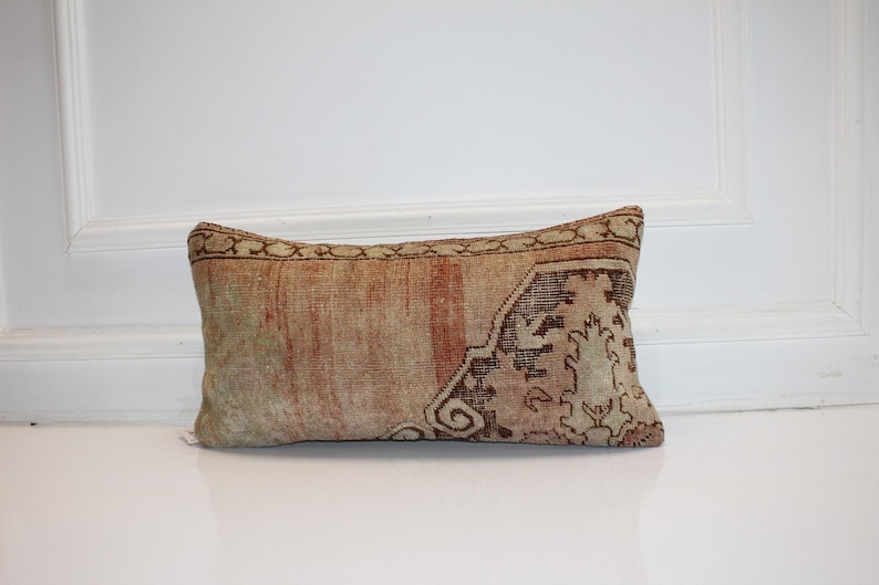 May include: A rectangular lumbar pillow featuring a faded, weathered textile cover with a muted color palette of beige, soft red, and brown. The fabric displays a worn, distressed texture with intricate, faded geometric and floral patterns.