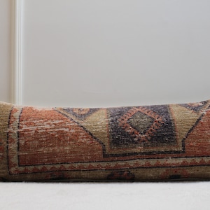 May include: A long, rectangular pillow with a colorful, geometric pattern. The pillow is made of a woven fabric with a distressed look. The colors in the pattern include red, orange, brown, and black.