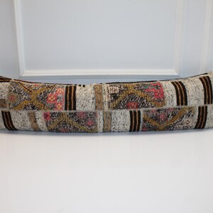 Puede incluir: Un largo cojín rectangular con un diseño de patchwork. El cojín presenta una mezcla de tonos neutros, incluyendo beige, marrón y crema, con toques de rosa y dorado. Rayas marrones verticales añaden un elemento de contraste. El cojín está sobre una superficie blanca.