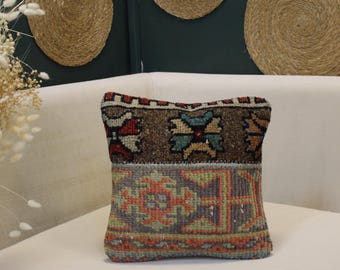 May include: A decorative pillow with a geometric pattern in shades of brown, red, green, and orange. The pillow is rectangular and appears to be made of woven fabric. The pillow is placed on a white surface.