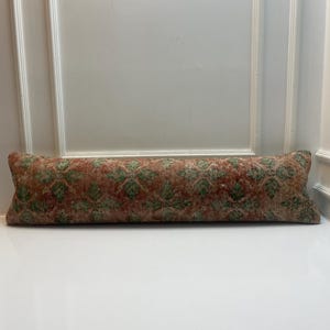Puede incluir: Un cojín cilíndrico largo y rectangular con una funda de tejido textil que presenta un patrón geométrico repetitivo en tonos naranja terracota, verde apagado y crema. La tela tiene una apariencia texturizada de inspiración vintage con detalles de tejido visibles.